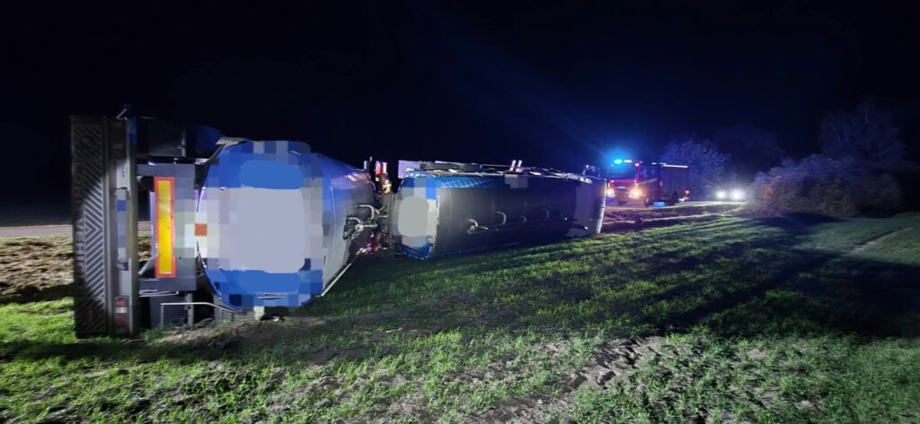 Przewrócona na bok cysterna leży na poboczu nocą, a w oddali świecą światła służb ratunkowych.
