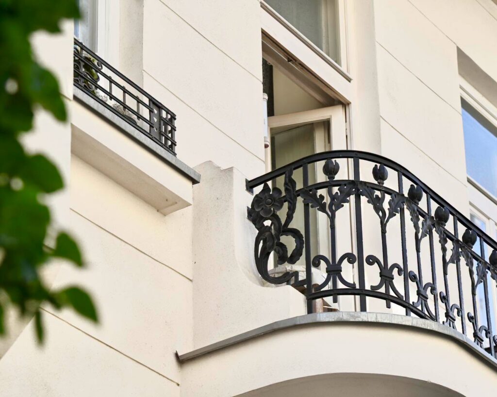 Ozdobny balkon z czarną kutą balustradą przy jasnej elewacji budynku i otwartym oknie.
