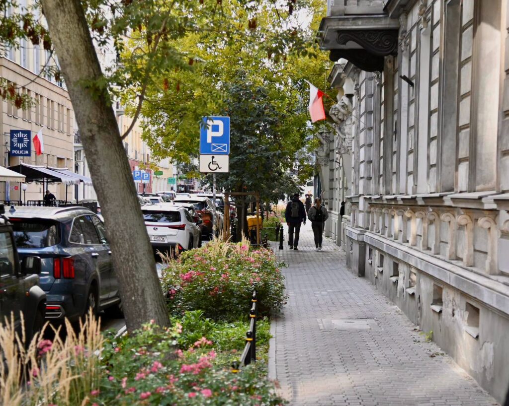 Dwie osoby idą chodnikiem wzdłuż zabytkowej kamienicy, obok zaparkowanych samochodów i znaku parkingu dla osób z
