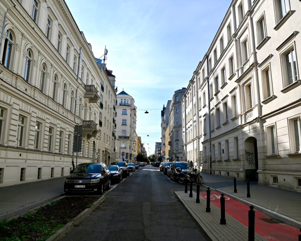 Wąska ulica z szeregiem jasnych kamienic, zaparkowanymi samochodami i czerwonym pasem rowerowym.