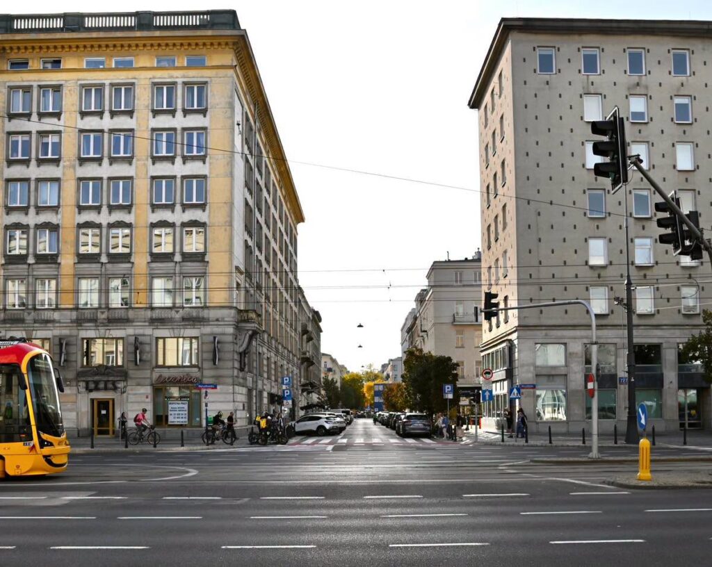 Skrzyżowanie z torami tramwajowymi między kamienicami, z samochodami, rowerzystami i pieszymi; z lewej strony tramwaj.