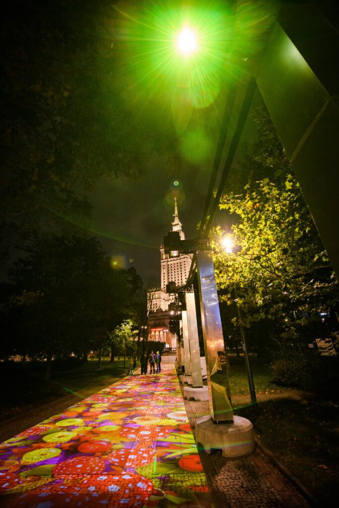Kolorowe iluminacje oświetlają parkową alejkę nocą, a w tle widać oświetlony wieżowiec i kilka osób.