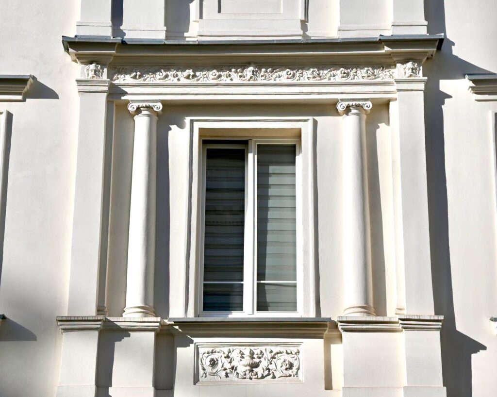 Wysokie okno z roletami w jasnej elewacji ozdobionej kolumnami i rzeźbionymi ornamentami.
