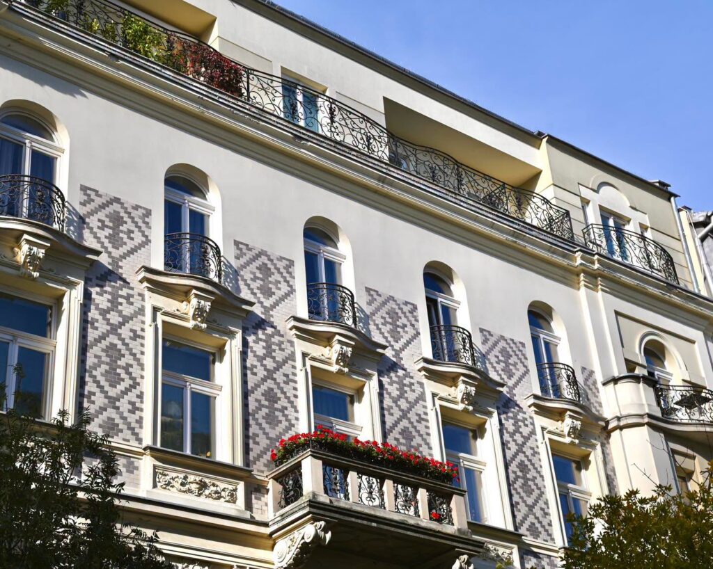 Jasna, wielopiętrowa kamienica z balkonami, kutymi balustradami i szaro-białą dekoracją elewacji pod błękitnym niebem.