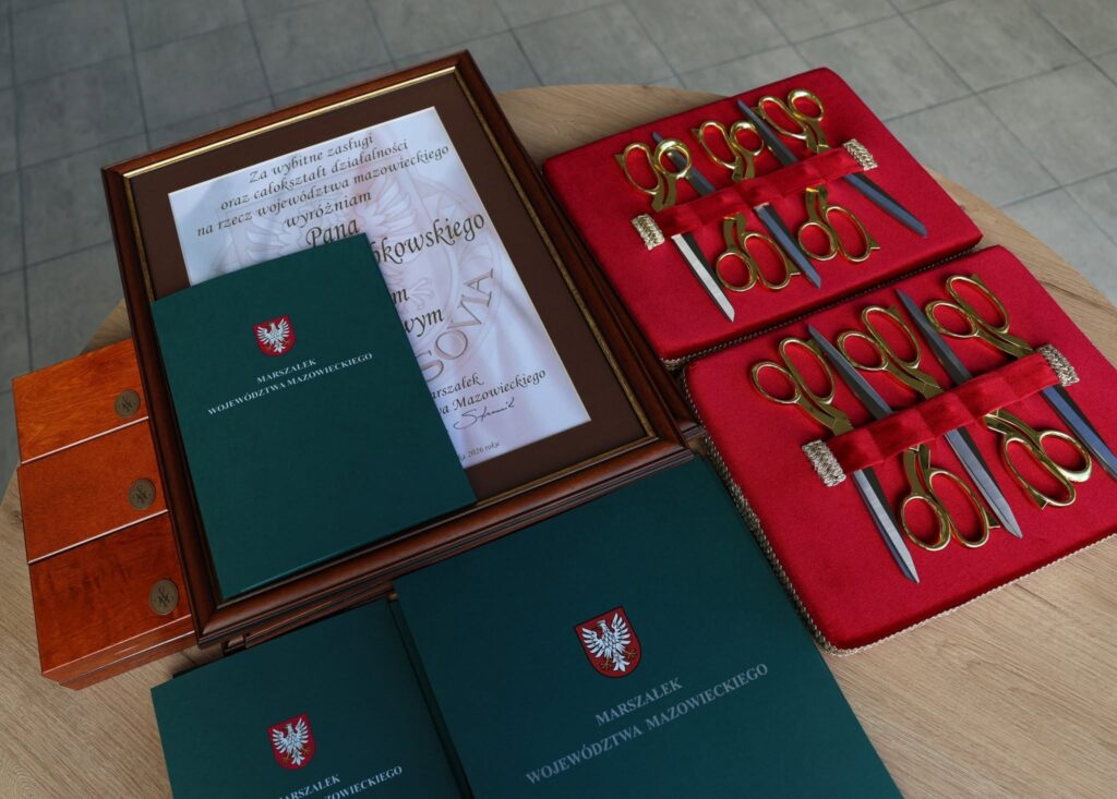 Zestawy nożyczek ceremonialnych, dyplom w ramie i zielone teczki z godłem leżą na drewnianym stole.