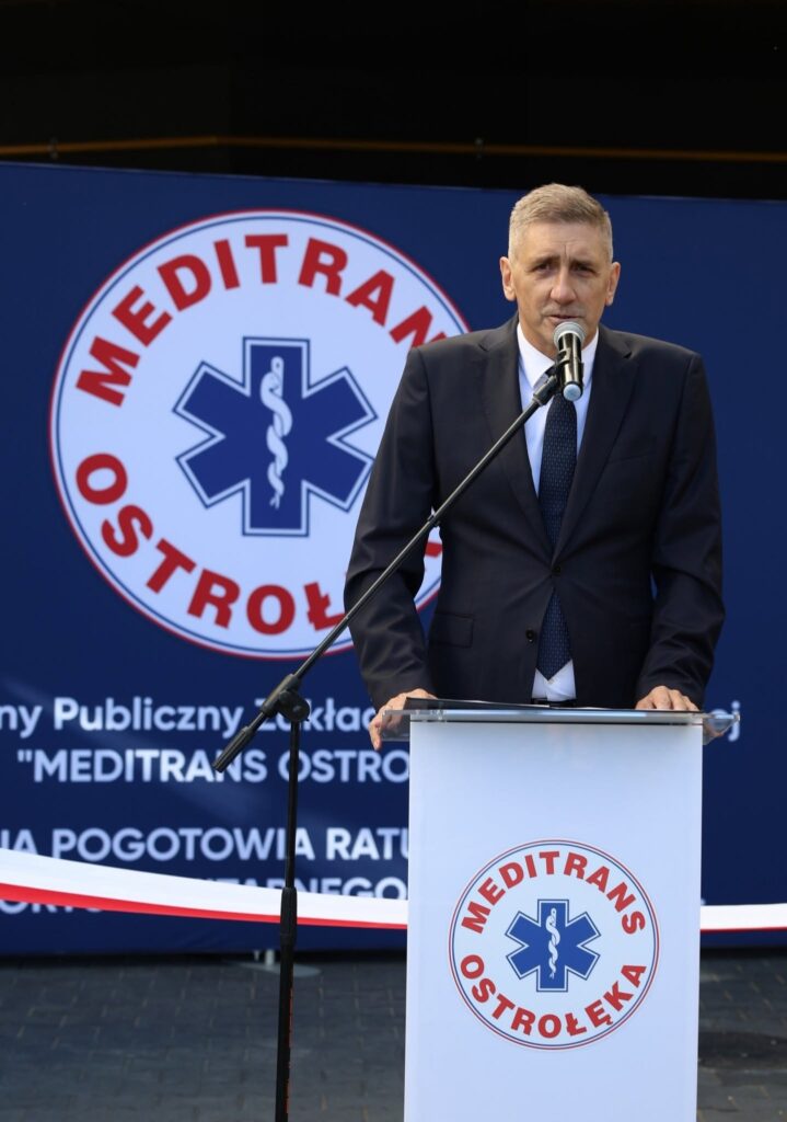 Mężczyzna w garniturze przemawia przy mównicy z logo „MEDITRANS OSTROŁĘKA”, na tle baneru z symbolem ratownictwa medycznego.