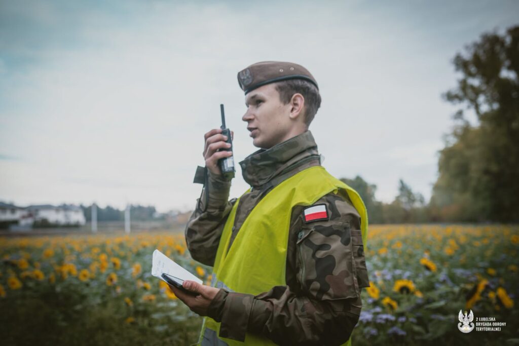 Żołnierz w kamuflażu i żółtej kamizelce rozmawia przez radiotelefon na łące z kwiatami, trzymając dokumenty.