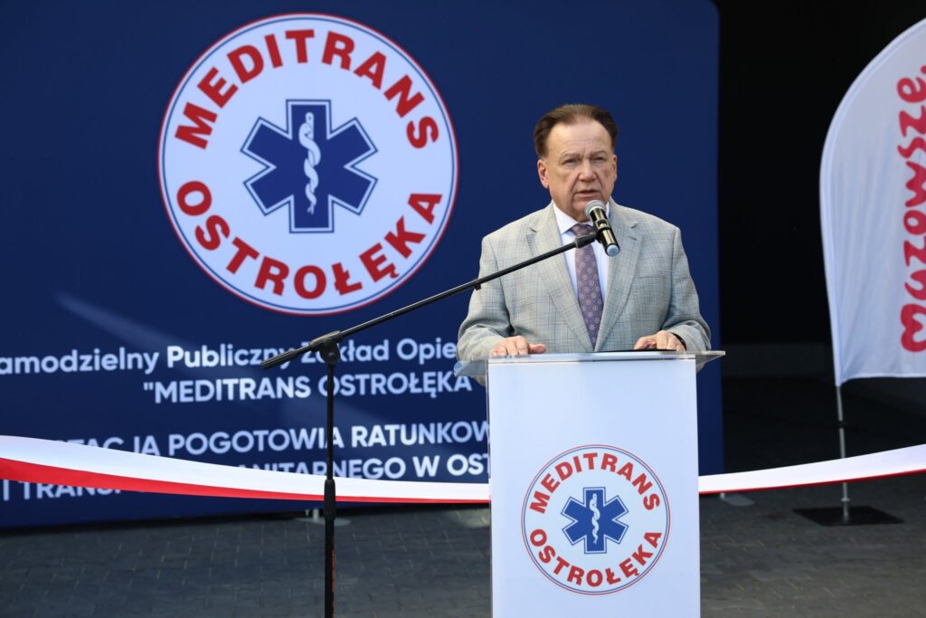 Mężczyzna w szarym garniturze przemawia przy mównicy z logo Meditrans Ostrołęka na tle baneru z tą samą nazwą.