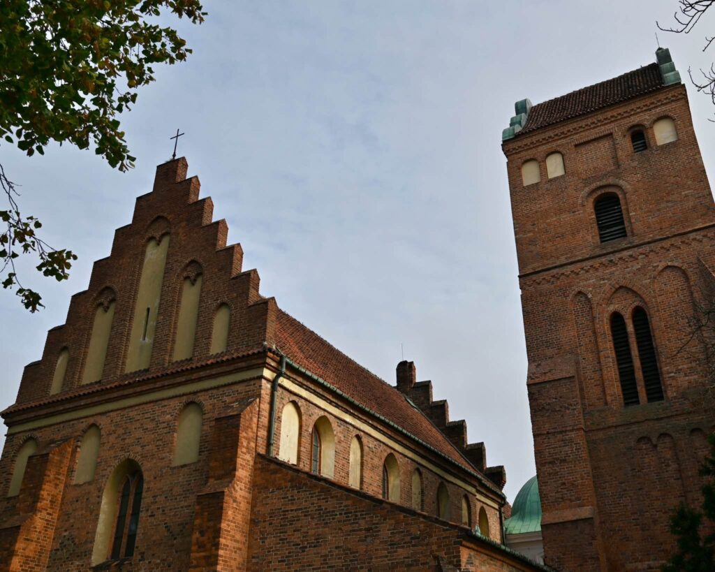 Ceglane zabudowania kościoła z wysoką wieżą i schodkowym szczytem widziane od dołu na tle nieba.