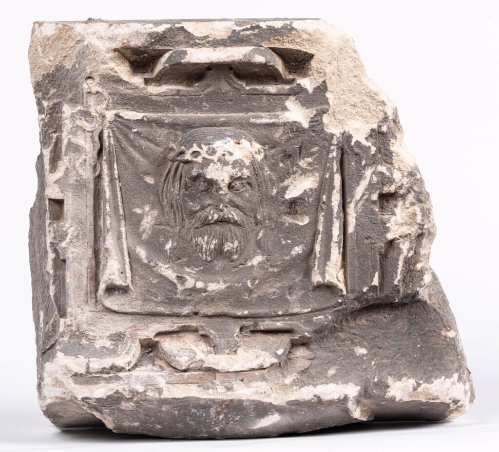 Uszkodzony kamienny relief z rzeźbioną twarzą brodatego mężczyzny, na jasnym tle.