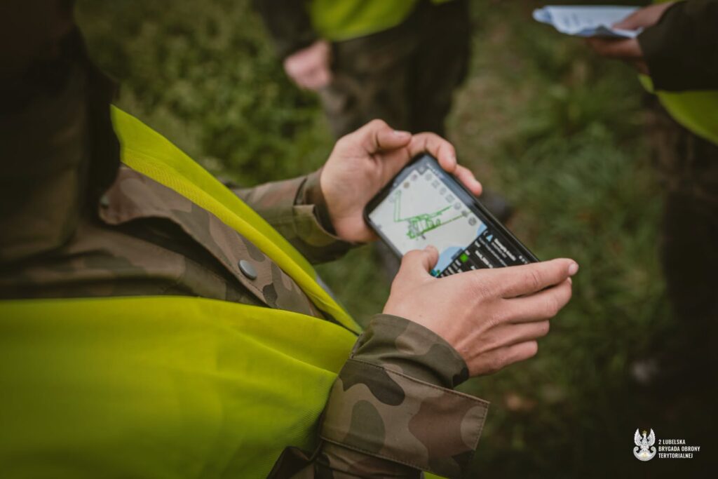 Osoba w mundurze i żółtej kamizelce sprawdza mapę na smartfonie w zalesionym terenie.