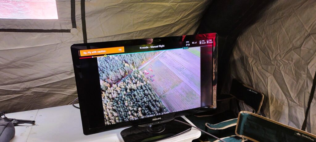 Monitor w namiocie wyświetla widok z lotu ptaka na las i pobliskie pola, obok leżą krzesła i sprzęt.