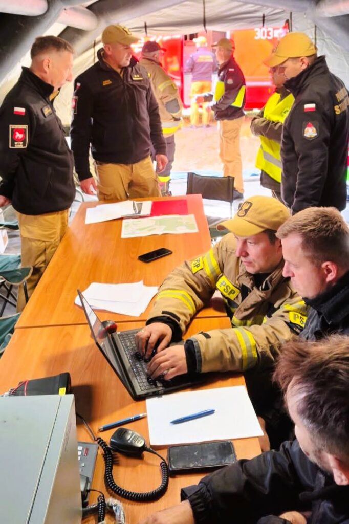 Strażacy i służby w namiocie operacyjnym omawiają dokumenty, a dwóch strażaków pracuje przy laptopie.