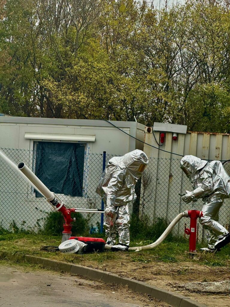 Dwóch strażaków w srebrnych kombinezonach ochronnych obsługuje węże przy hydrantach na ogrodzonym terenie przemysłowym.