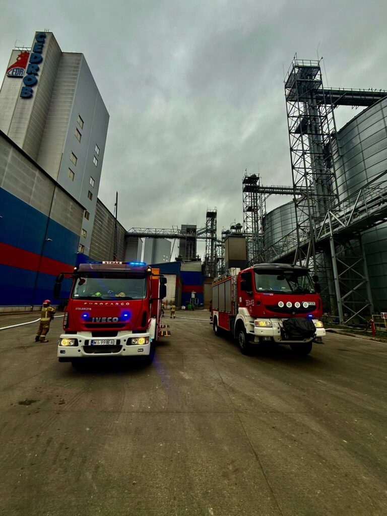 Dwa czerwone wozy strażackie stoją na terenie zakładu przemysłowego, wśród silosów i metalowych konstrukcji.
