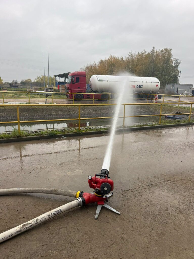 Strażacka prądownica wyrzuca strumień wody w kierunku cysterny z napisem „BOB GAZ” na terenie zakładu przemysłowego.
