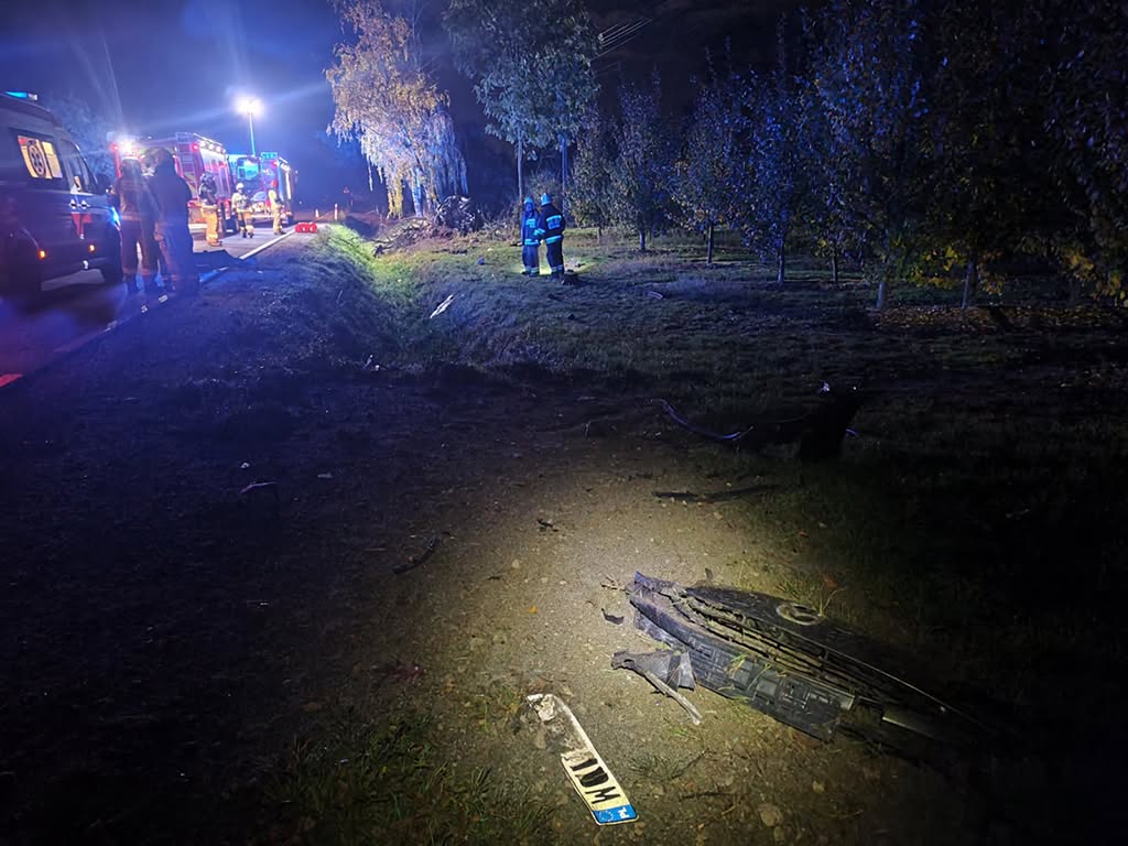 Strażacy pracują nocą przy drodze obok uszkodzonego samochodu, a na ziemi leżą oderwane elementy pojazdu.