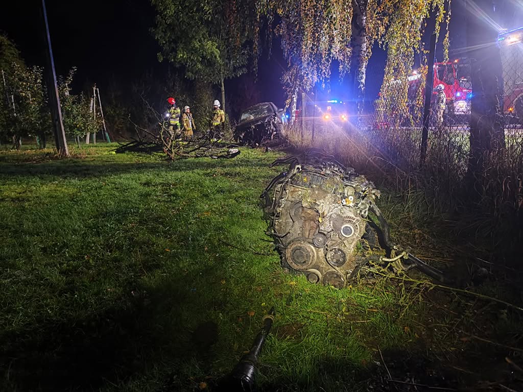 Silnik leży na trawie obok rozbitego samochodu, a strażacy pracują nocą przy oświetlonych pojazdach ratunkowych.