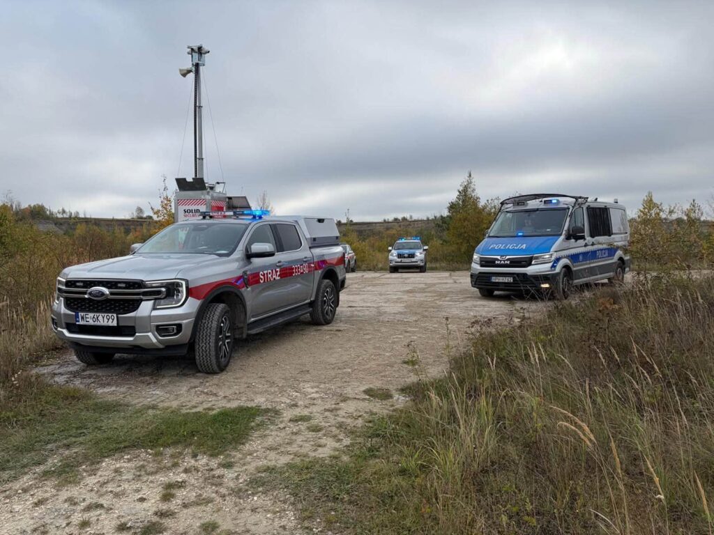 Samochód straży pożarnej i dwa radiowozy stoją na polnej drodze wśród zarośli przy pochmurnej pogodzie.