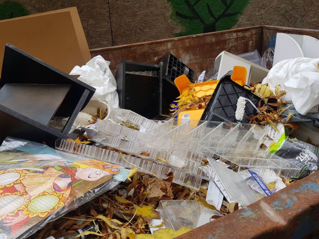 Porzucone plastikowe pojemniki, fragmenty mebli, worki i liście leżą na stercie śmieci w metalowym kontenerze.