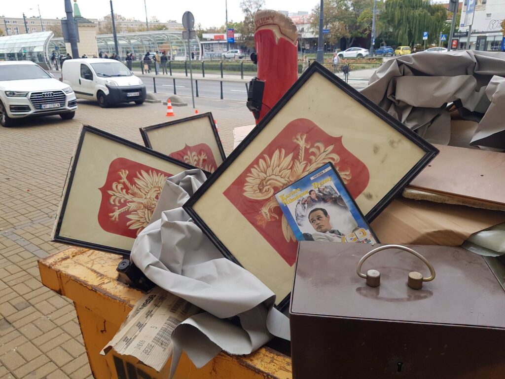 Oprawione polskie godła i inne przedmioty leżą wśród porzuconych mebli i tkanin na ulicznym śmietniku.