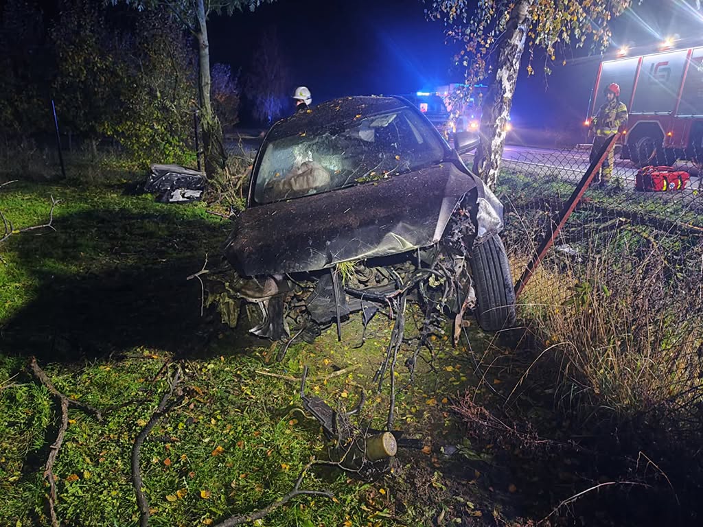 Rozbita maźda z mocno uszkodzonym przodem stoi przy ogrodzeniu, obok strażacy i wóz strażacki nocą.