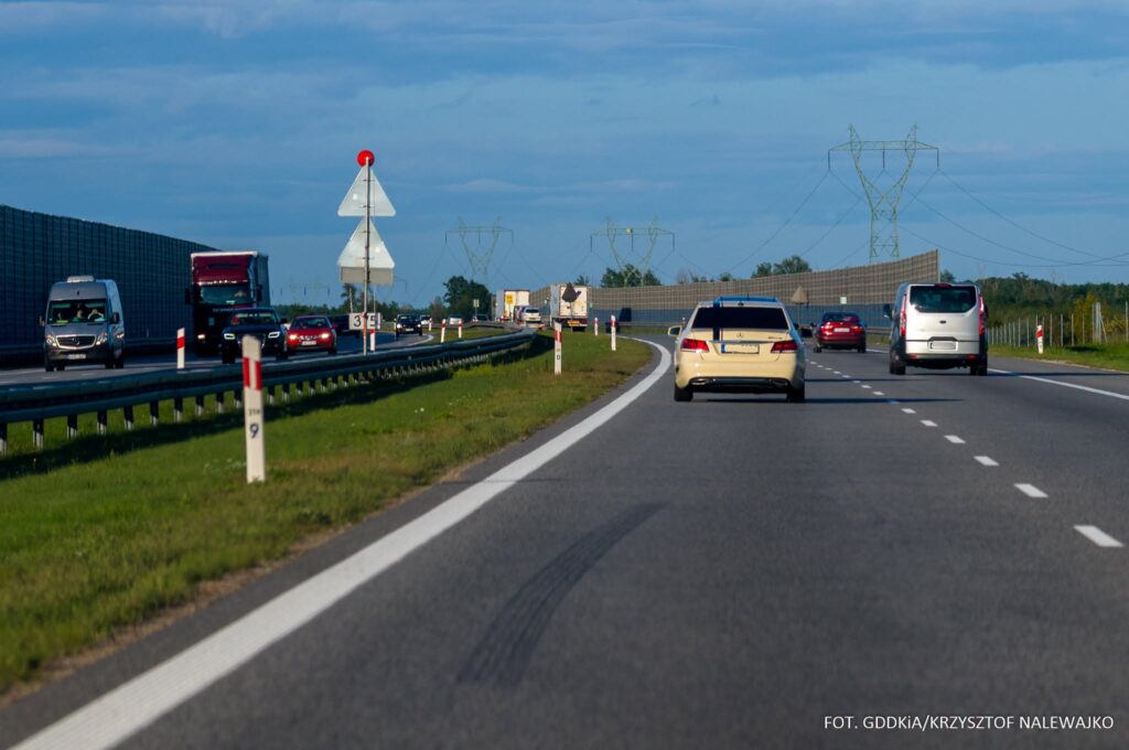 Samochody i ciężarówki jadą wielopasmową autostradą z barierami energochłonnymi i ekranami akustycznymi.