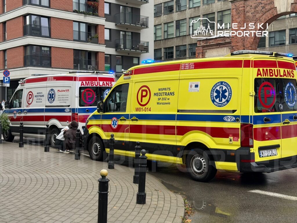 Dwie karetki pogotowia zaparkowane przy chodniku przed blokami mieszkalnymi; obok jednej klęczy ratownik.