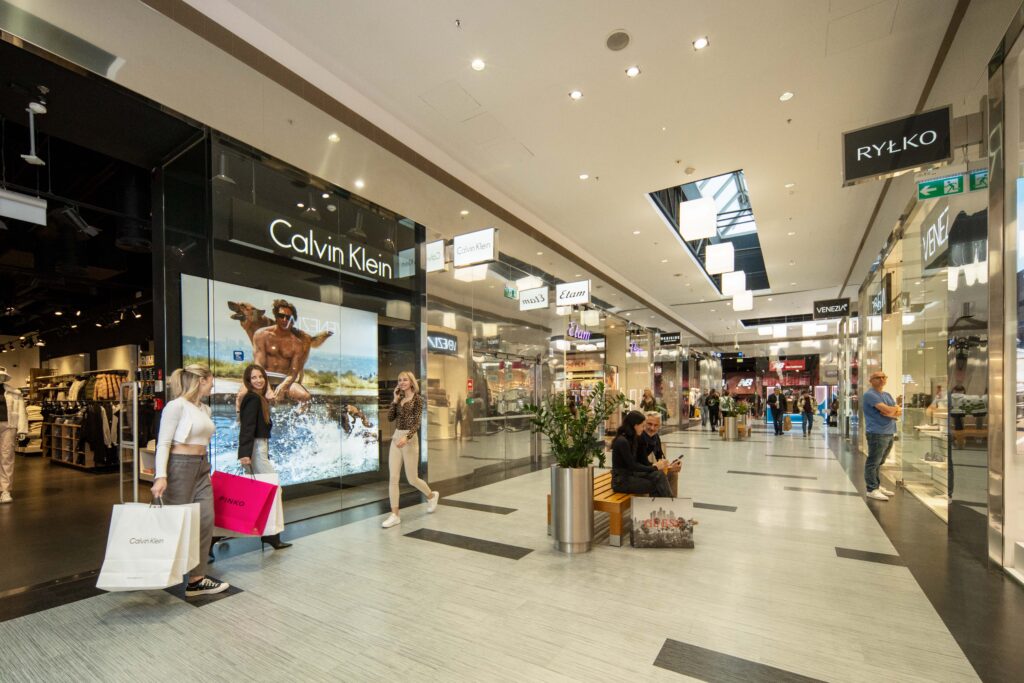 Klienci z torbami zakupowymi przechodzą pasażem centrum handlowego obok sklepów Calvin Klein i RYŁKO.