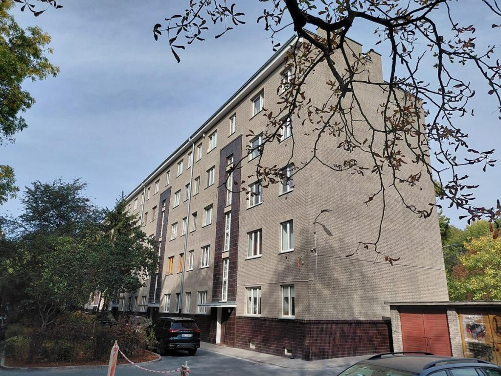Odnowiona, kilkupiętrowa kamienica z jasną elewacją i czerwonymi bramami, otoczona drzewami i zaparkowanymi samochodami.