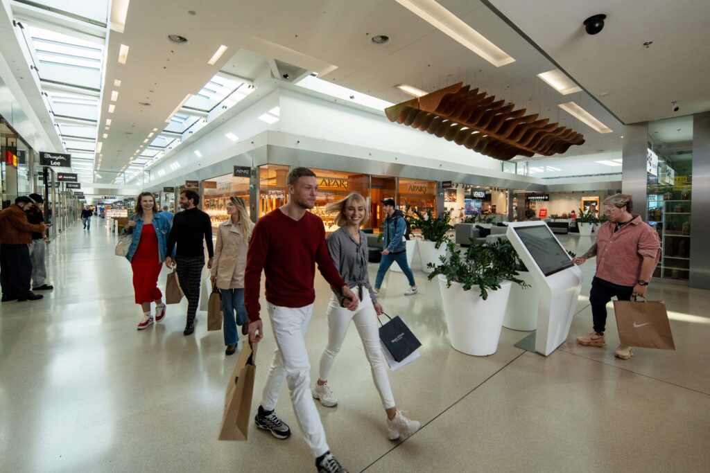 Klienci robią zakupy w przestronnym centrum handlowym, mijając sklepy i rośliny doniczkowe.