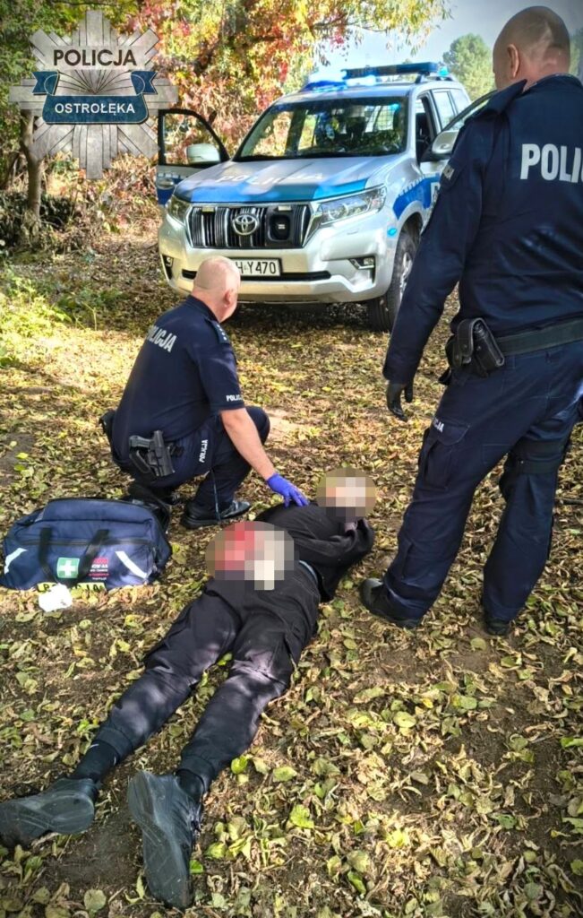 Policjanci udzielają pomocy leżącej osobie obok radiowozu z napisem „Ostrołęka” i torby medycznej.