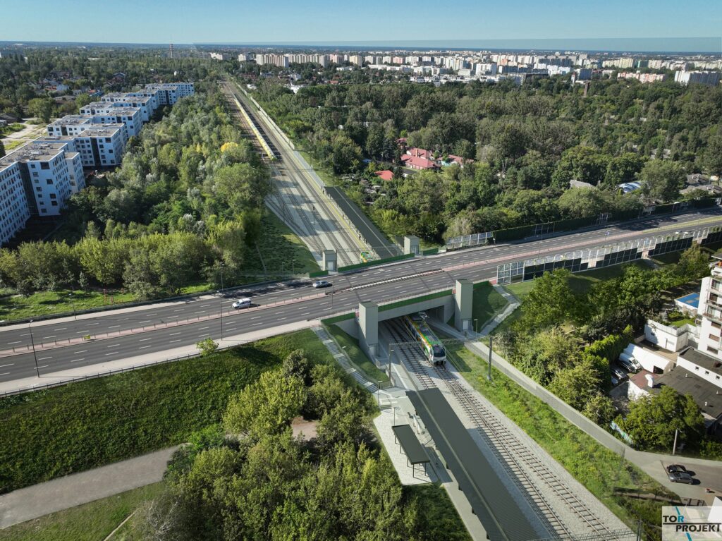 Widok z lotu ptaka na tory kolejowe biegnące pod wiaduktem drogowym, otoczone zielenią i zabudową mieszkaniową.
