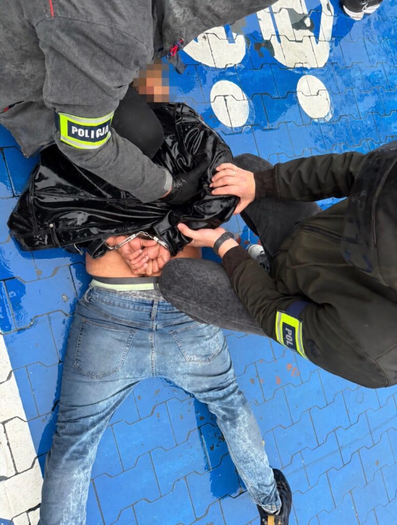 Policjanci obezwładniają i zakuwają w kajdanki mężczyznę leżącego na chodniku z niebieskiej kostki brukowej.