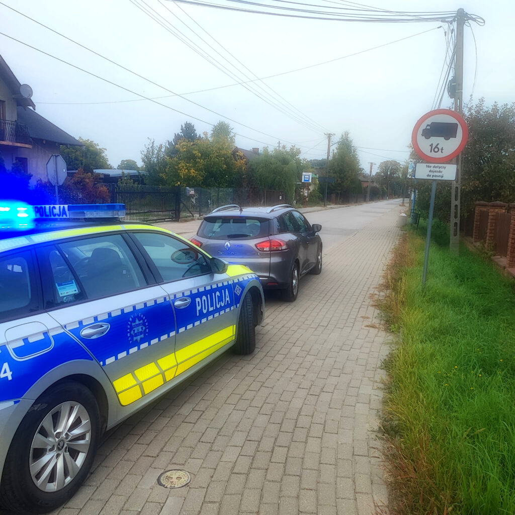 Oznakowany radiowóz policyjny stoi za szarym samochodem na brukowanej ulicy, przy znaku zakazu wjazdu ciężarówek powyżej 16