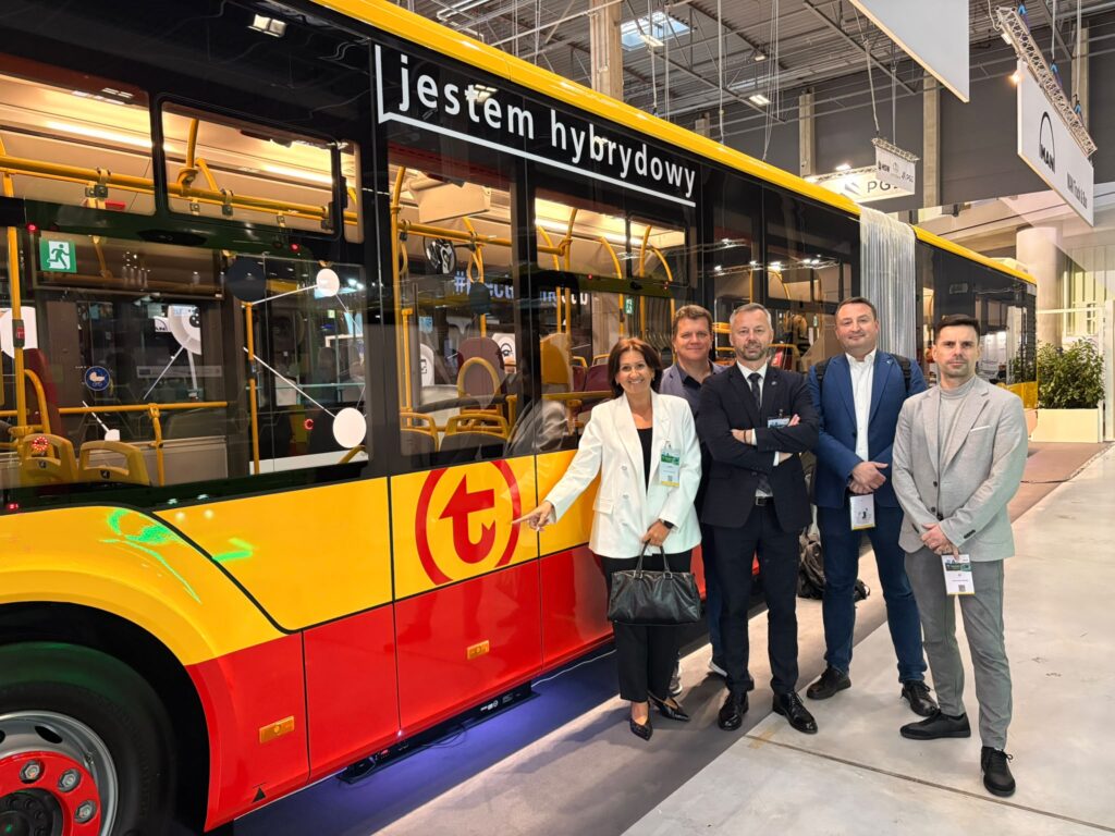 Pięć osób pozuje obok żółto-czerwonego autobusu z napisem „jestem hybrydowy” na hali wystawowej.