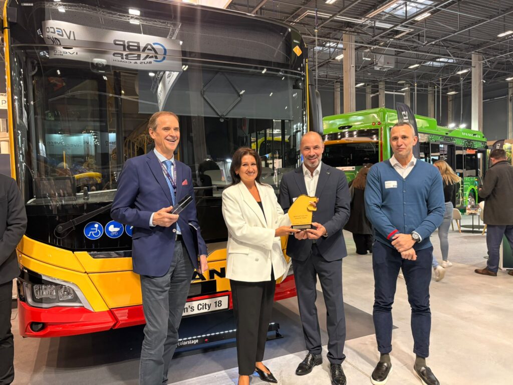 Czworo osób prezentuje żółtą statuetkę przed żółto-czarnym autobusem w hali wystawowej.