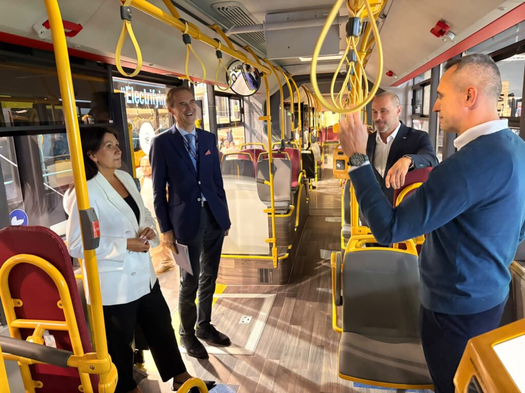 Cztery osoby rozmawiają we wnętrzu autobusu z żółtymi poręczami i czerwonymi siedzeniami.