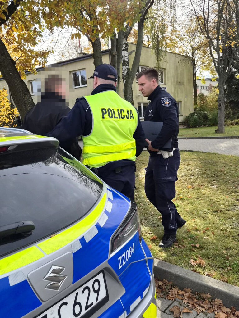 Policjanci rozmawiają z zamazanym mężczyzną obok oznakowanego radiowozu, na tle budynku i jesiennych drzew.