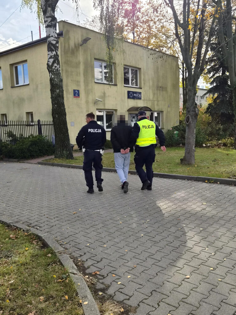 Dwóch policjantów prowadzi zatrzymanego mężczyznę przed budynkiem komisariatu policji.