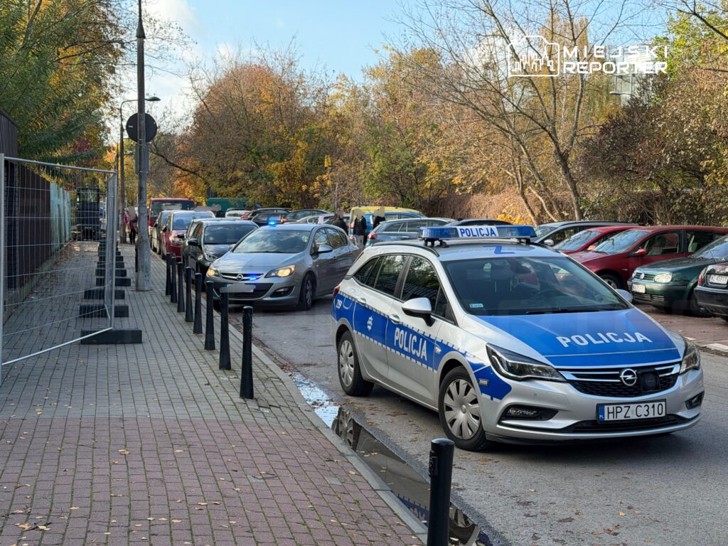 Radiowóz policyjny stoi na ulicy, wśród zaparkowanych samochodów i drzew w jesiennych barwach.