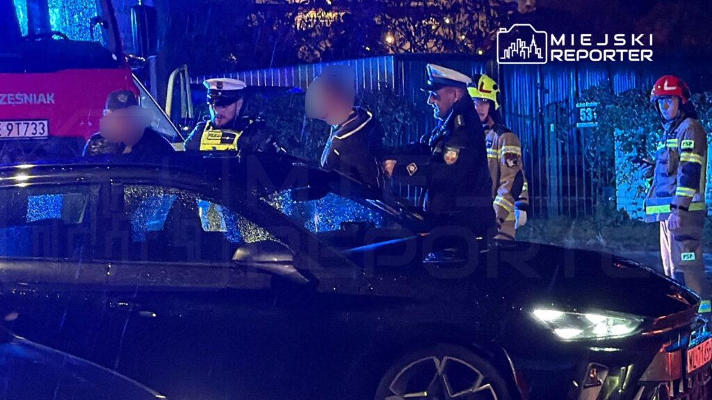 Policjanci i strażacy przy uszkodzonym czarnym samochodzie na nocnym miejscu zdarzenia, oświetlonym niebieskimi światłami