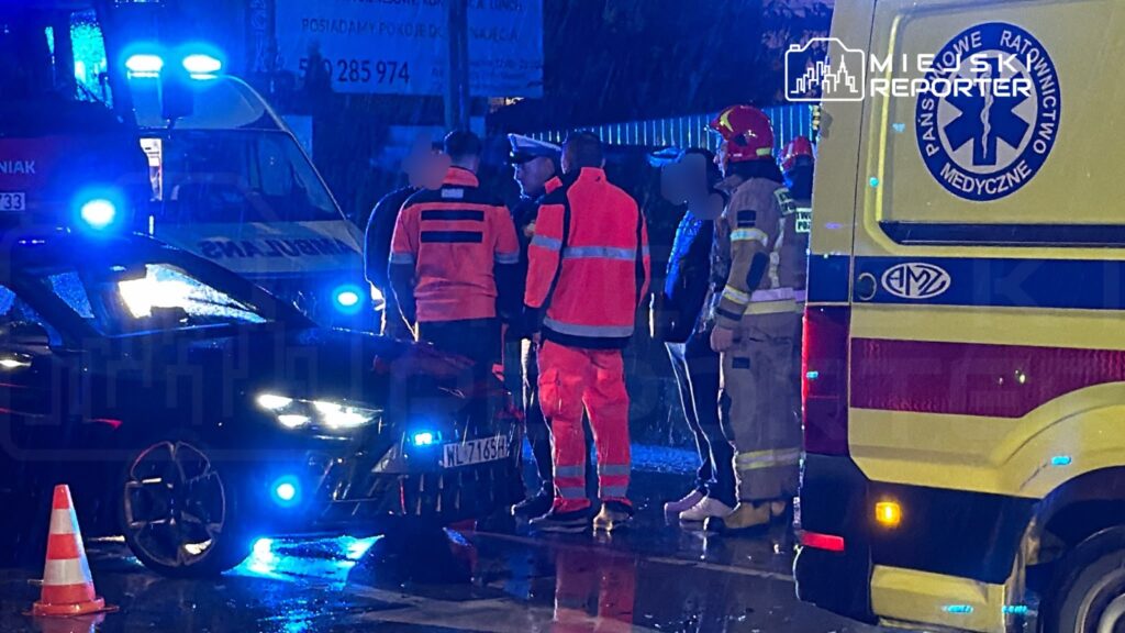Ratownicy medyczni i strażak stoją przy samochodzie na mokrej ulicy, obok karetki z włączonymi niebieskimi światłami.