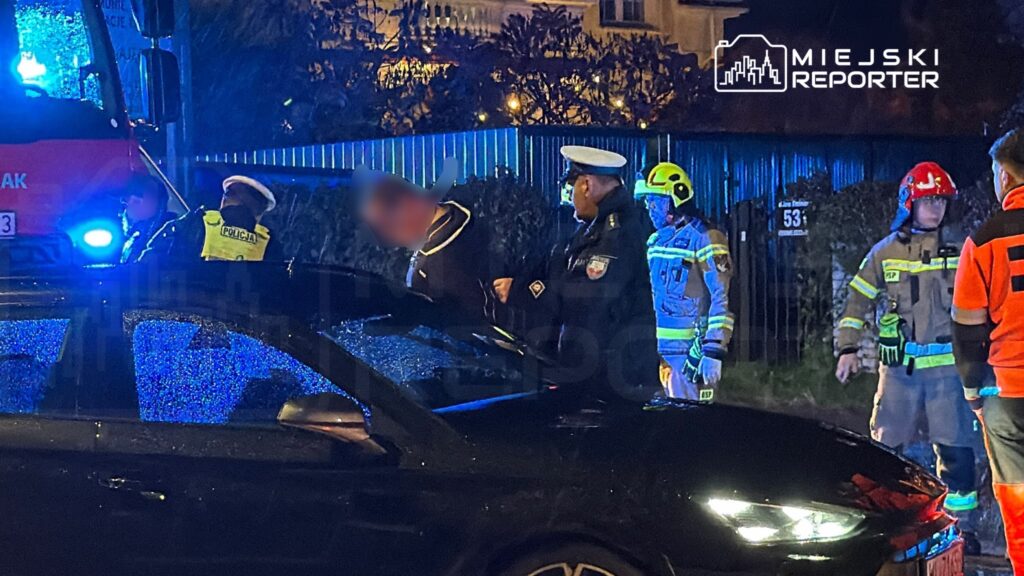 Policjanci i strażacy stoją przy czarnym samochodzie podczas nocnej akcji, obok radiowozu z włączonymi światłami.