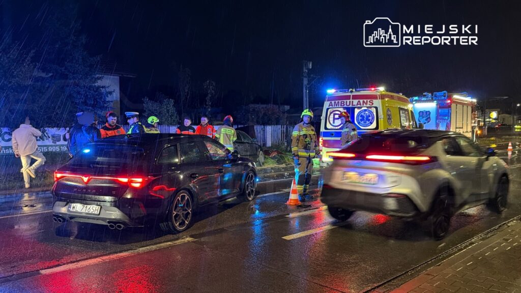 Ratownicy przy ambulansie i samochodzie po kolizji na mokrej, oświetlonej niebieskimi światłami ulicy nocą.