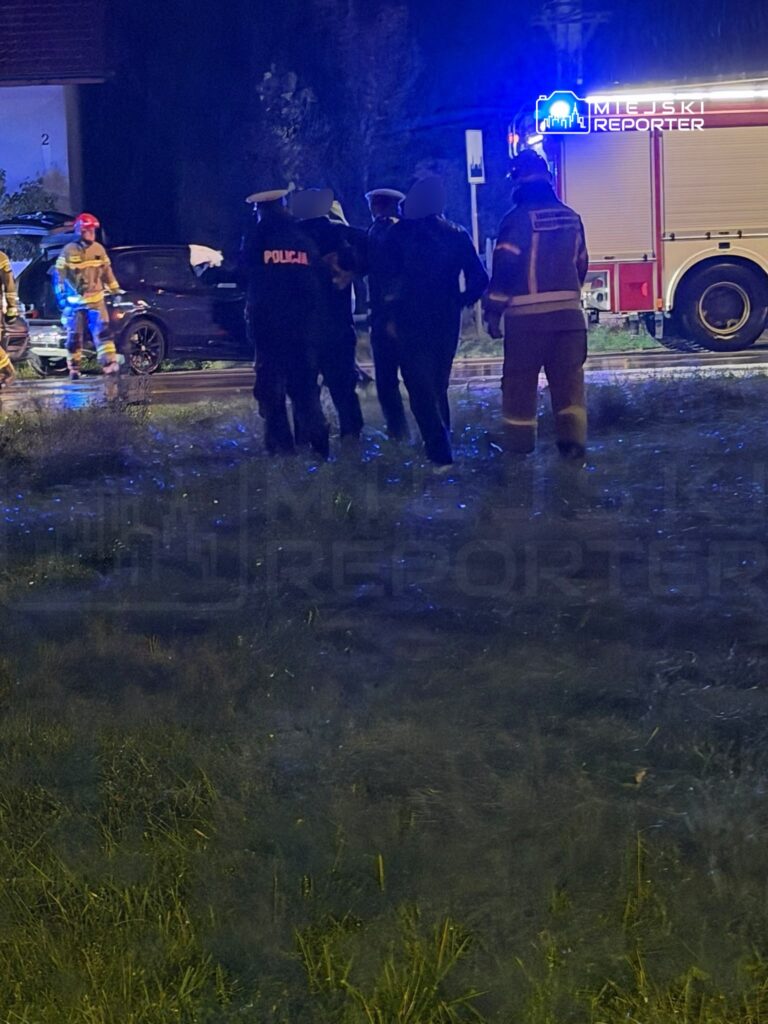 Policjanci i strażacy stoją przy samochodzie obok wozu strażackiego, na zalanej wodą ulicy nocą.
