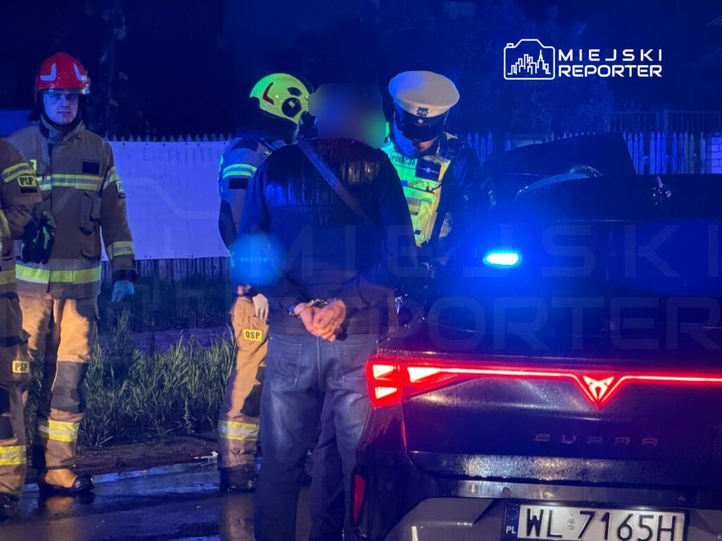Strażacy w odblaskowych ubraniach stoją obok mężczyzny z rękami za plecami i zaparkowanego auta nocą.