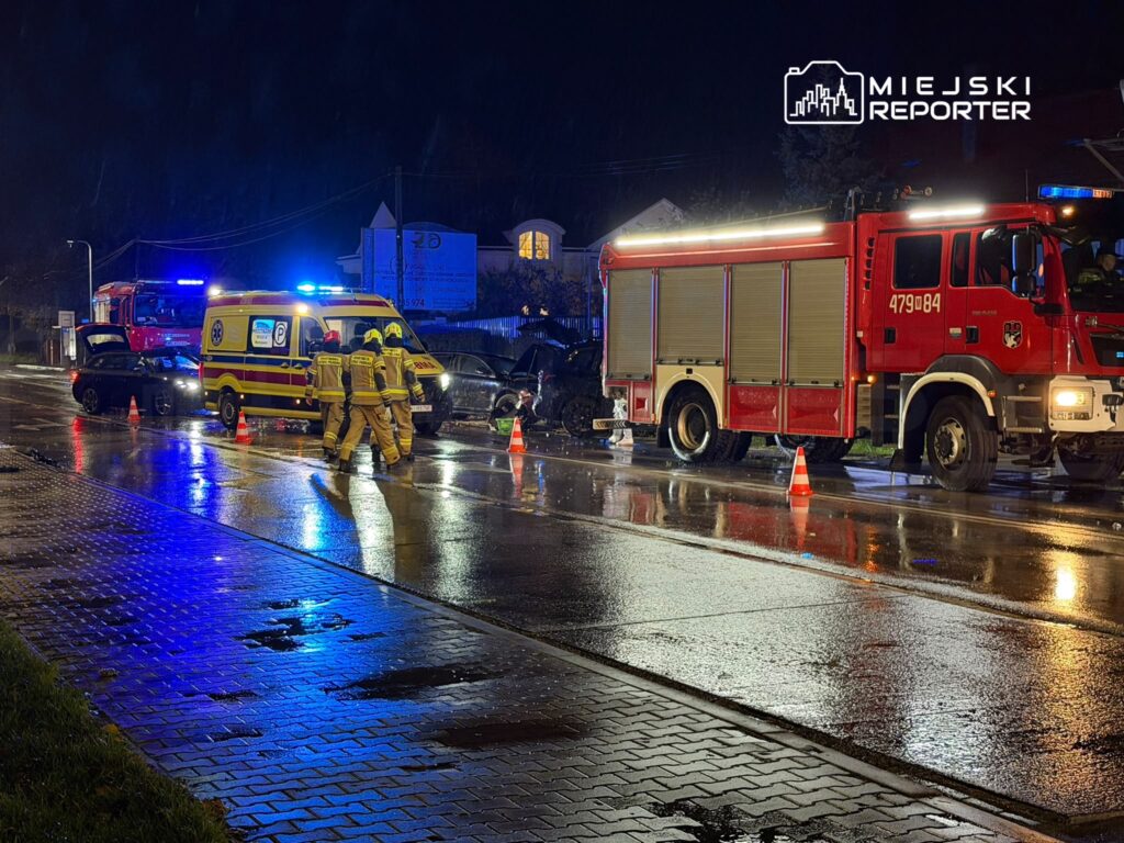 Strażacy stoją przy wozie strażackim i karetce na mokrej, oświetlonej drodze nocą.