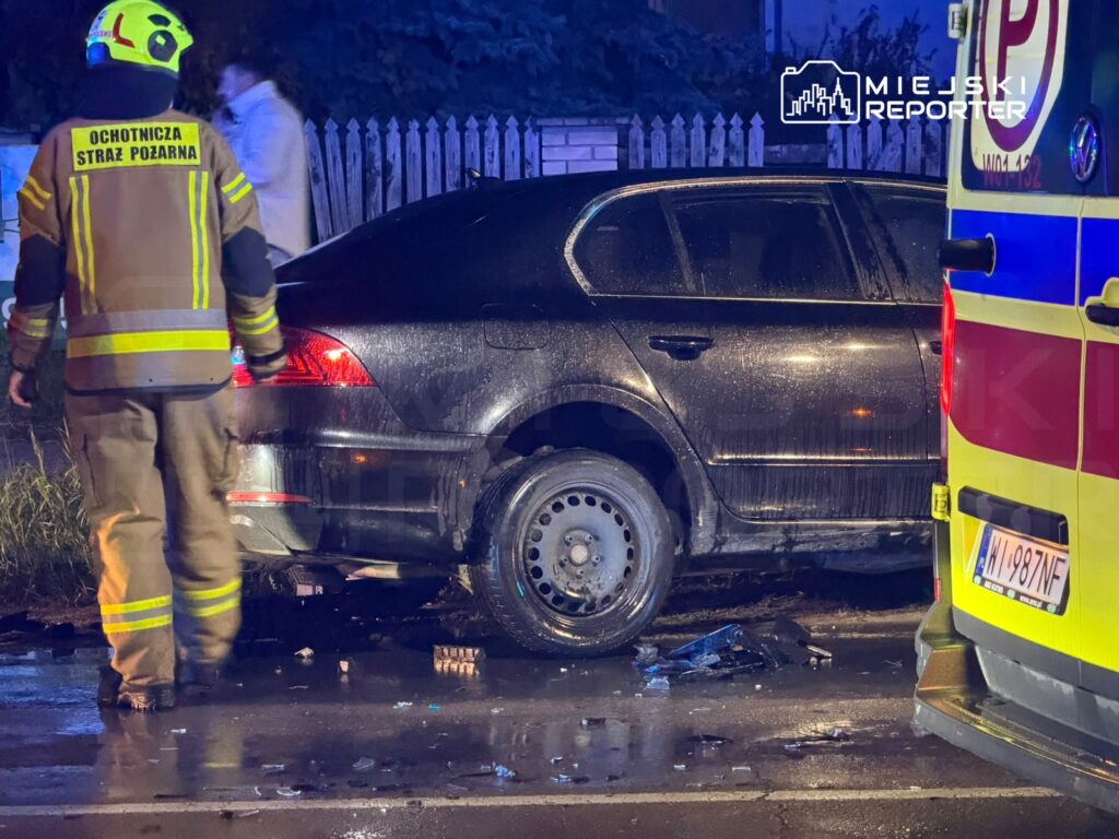 Strażak ochotniczej straży pożarnej stoi przy uszkodzonym samochodzie osobowym obok karetki, na mokrej jezdni nocą.