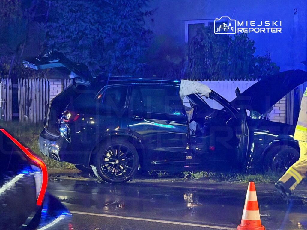Czarny samochód po zderzeniu stoi przy ogrodzeniu, z otwartymi drzwiami i uszkodzoną karoserią na mokrej jezdni.